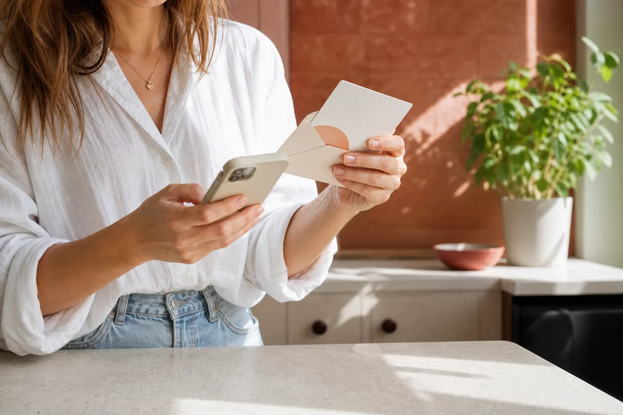 A woman checking her phone while holding a spa gift card and envelope